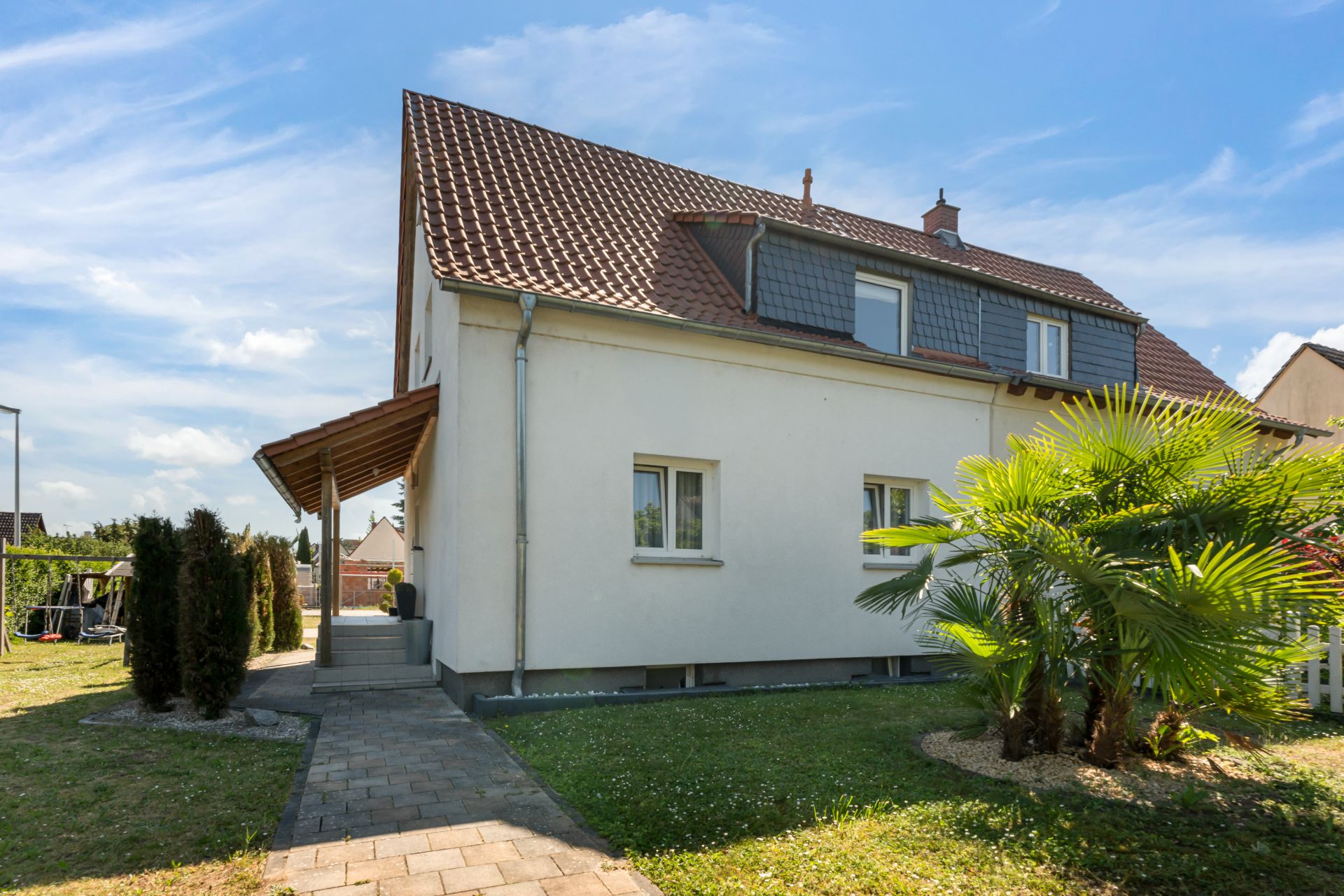 Zweistöckiges Wohnhaus mit rotem Dach, Garten, Palmen und gepflastertem Gehweg.
