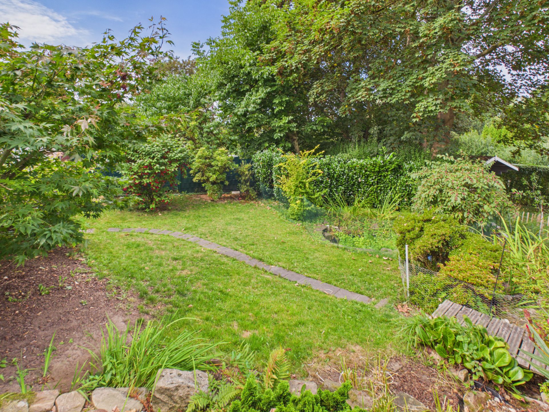 Grüner Garten mit Rasenfläche, Steinweg, Büschen und Bäumen.