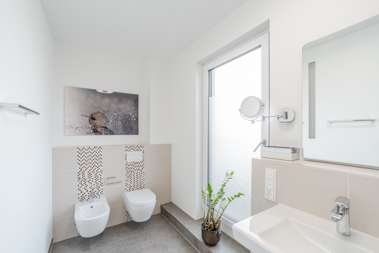 Helles, modernes Badezimmer mit Bidet, WC, großem Fenster und einer Pflanze auf dem Boden.
