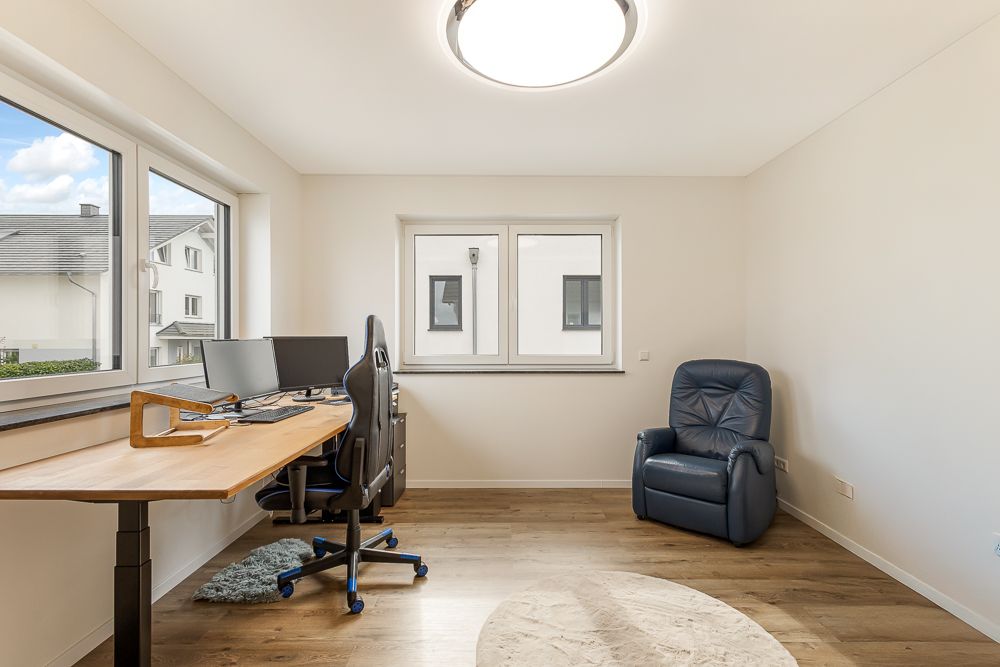 Helles Arbeitszimmer mit Schreibtisch, Bürostuhl und Sessel vor zwei großen Fenstern.