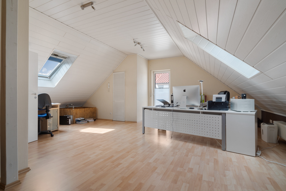 Helles Dachzimmer mit Schreibtischen, Stühlen und Computern, zwei Dachfenstern und Laminatboden.