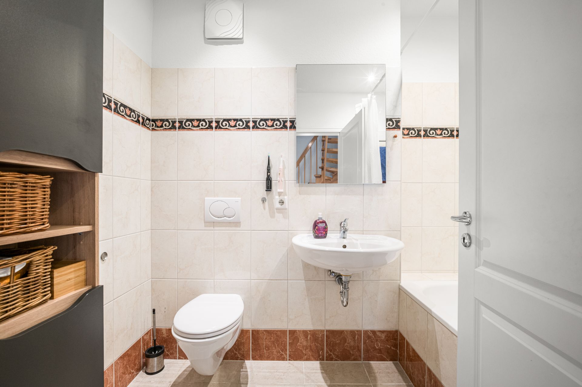 Helles Badezimmer mit WC, Waschbecken, Spiegel, Badewanne und Regal mit Körben.