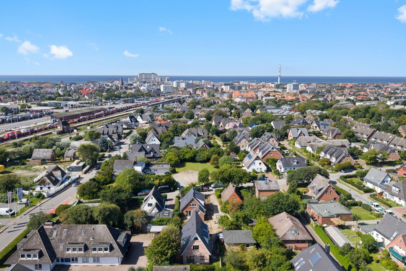 Luftaufnahme eines Wohngebiets mit vielen Häusern, Bahngleisen und Blick auf das Meer im Hintergrund.