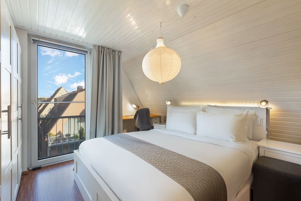 Helles Schlafzimmer mit Doppelbett, Schreibtisch, großer Fensterfront und weißer Holzdecke.