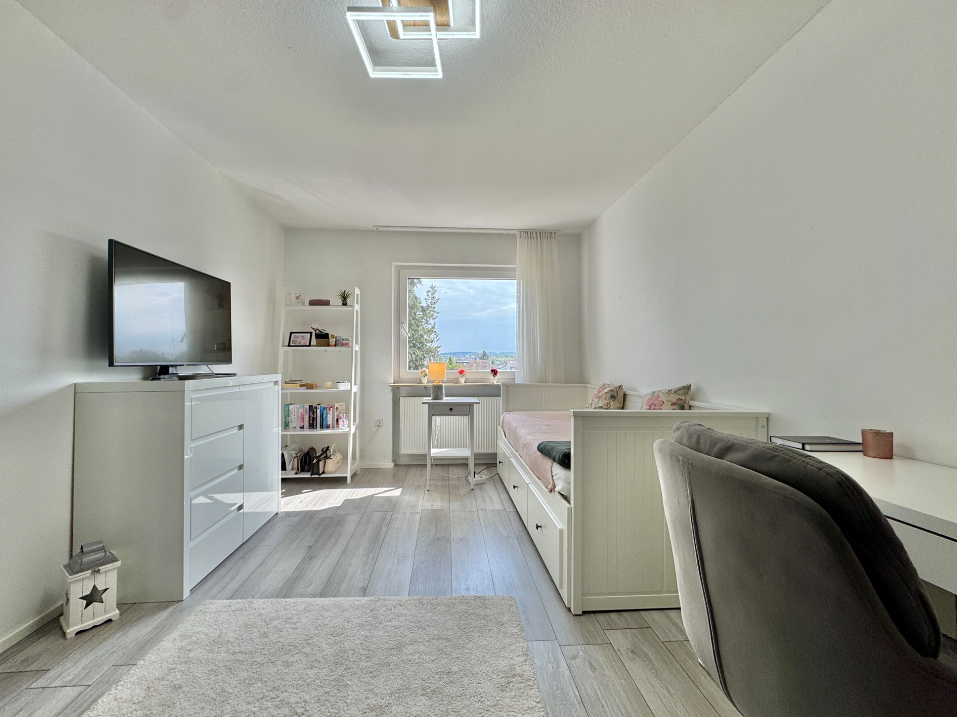 Helles Schlafzimmer mit TV, Bett, Schreibtisch, Regal und Fenster mit Blick nach draußen.