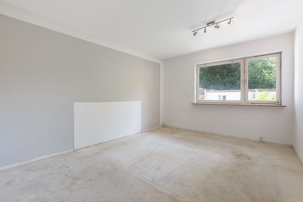 Leeres Zimmer mit hellem Teppich, großem Fenster und Deckenleuchte.