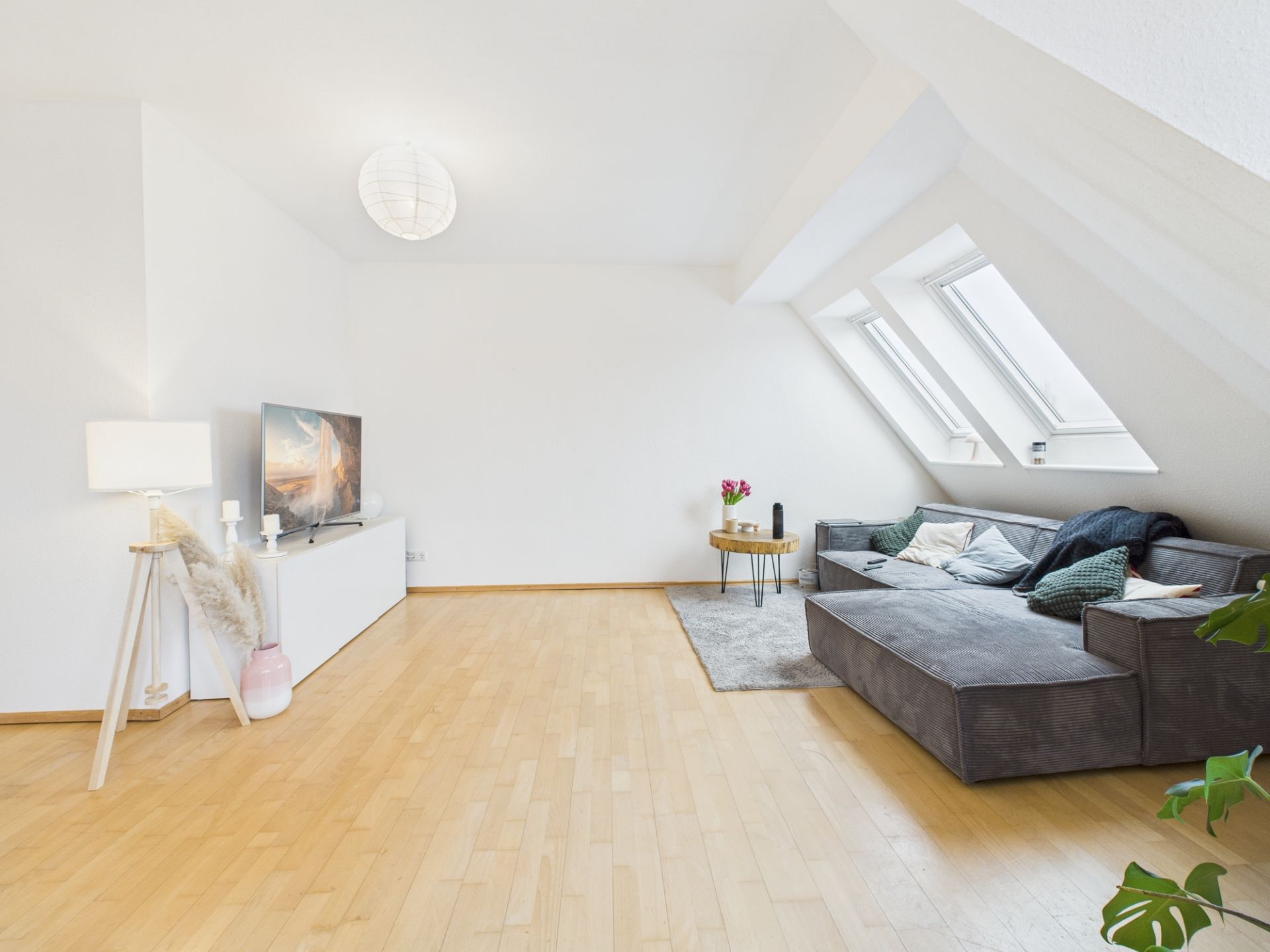 Helles Wohnzimmer mit Ecksofa, Teppich, TV, Stehlampe und Dachfenstern.