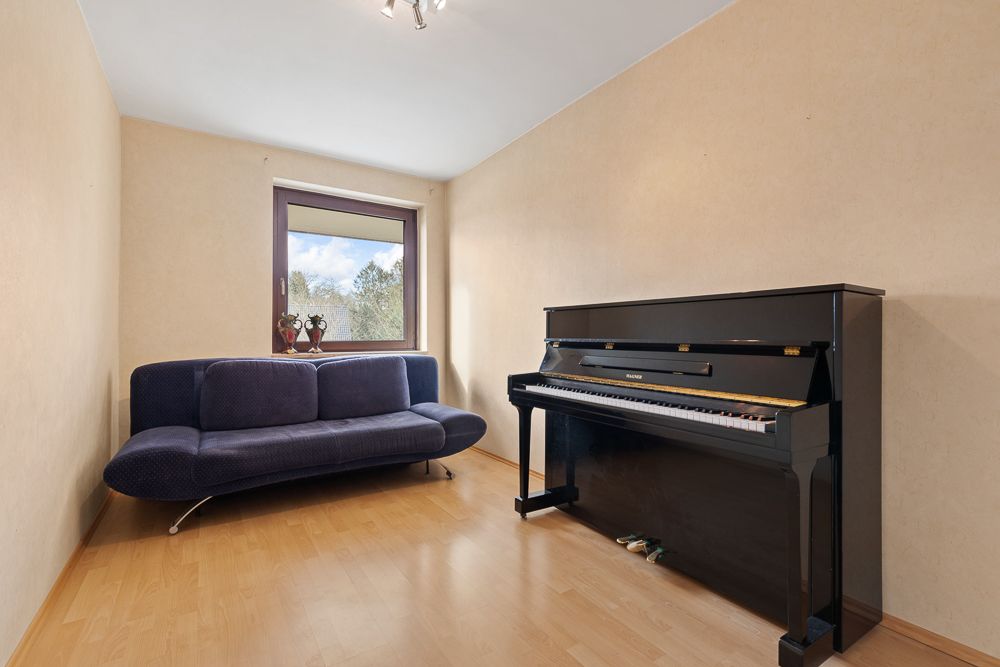 Schmales Zimmer mit dunkelblauem Sofa, schwarzem Klavier und Fenster mit Blick ins Grüne.