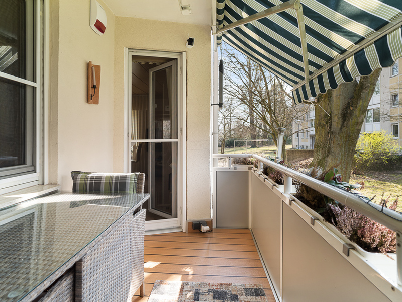 Möblierter Balkon mit Markise, Glastisch und Blick auf Bäume und Nachbargebäude.