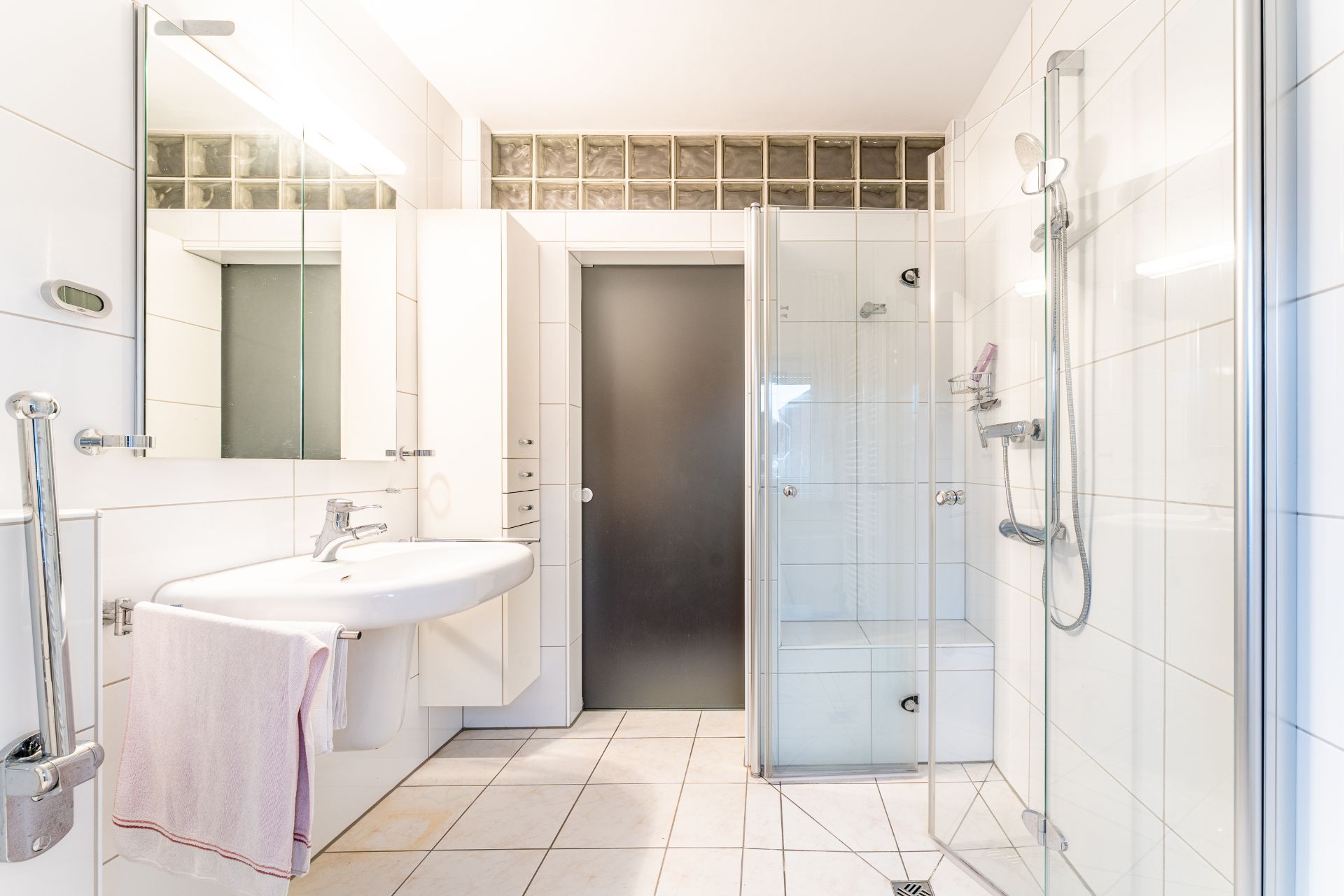 Helles Badezimmer mit Waschbecken, Spiegel, Dusche und weiß gefliesten Wänden.