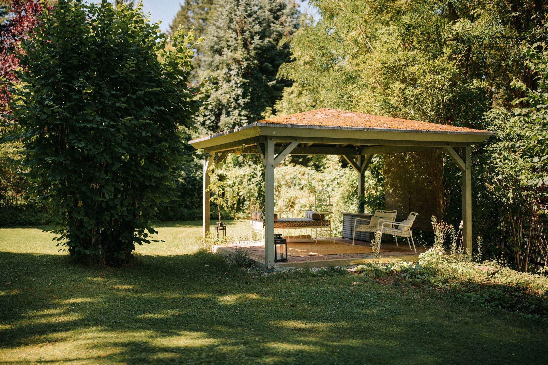 Überdachter Pavillon mit Sitzmöbeln im Garten, umgeben von Bäumen und Rasen.