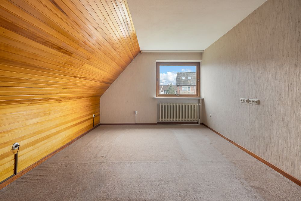 Leeres Zimmer mit Teppichboden, Holzverkleidung an der Schräge und Fenster mit Heizkörper.