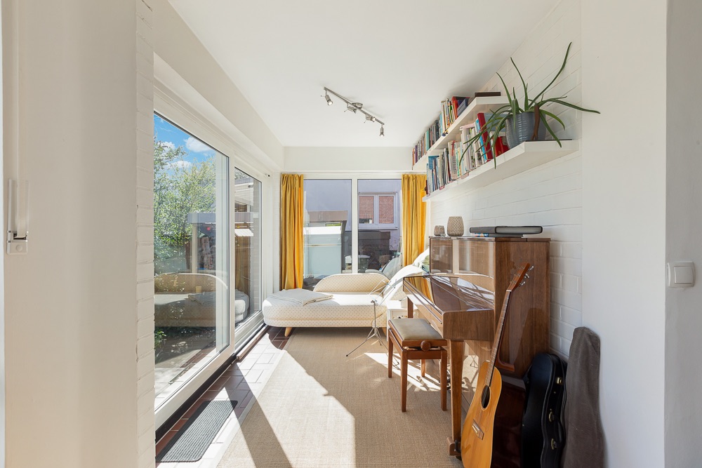 Heller Raum mit großem Fenster, Sofa, Klavier, Gitarre und Regal mit Pflanzen und Büchern.