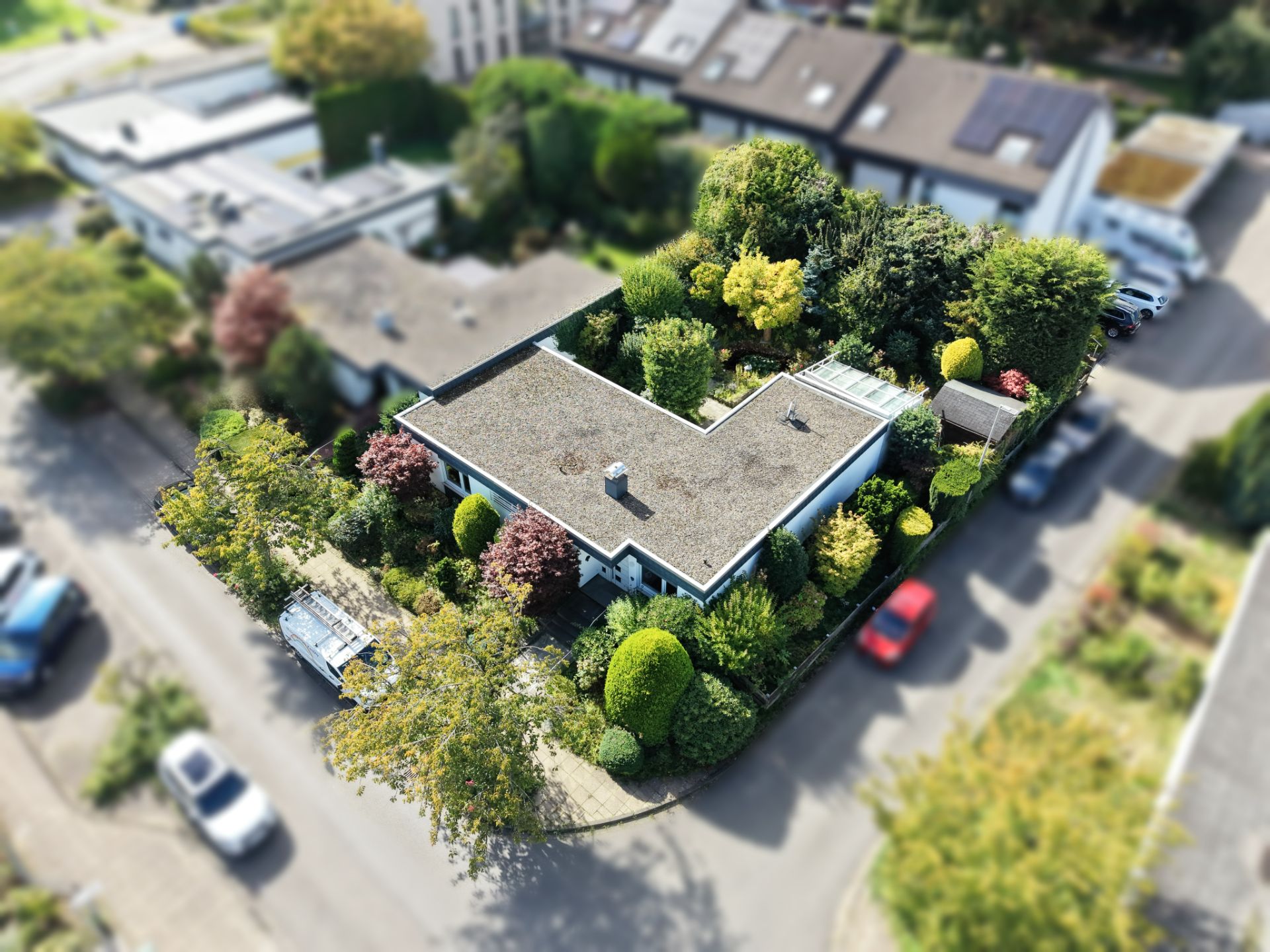 Luftaufnahme eines Hauses mit Flachdach und großem, grünem Garten an einer Straßenecke.