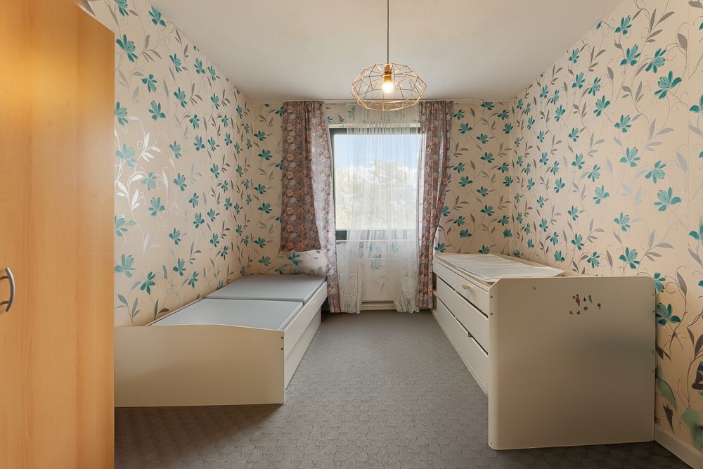 Kinderzimmer mit Bett, Kommode und floraler Tapete, Fenster mit Gardinen in der Mitte.