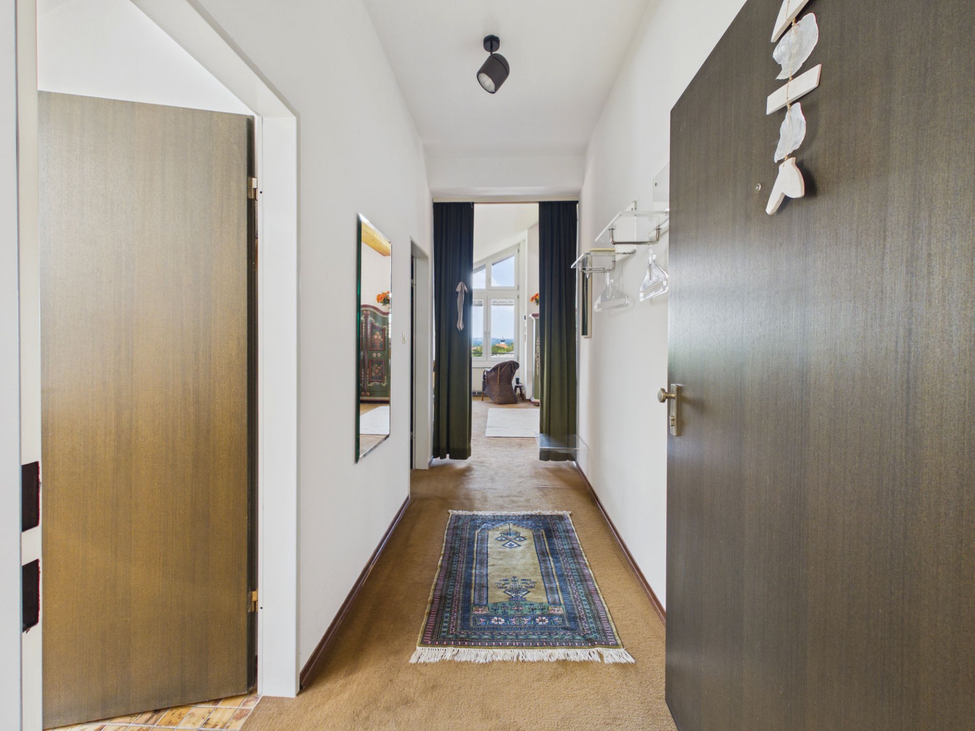 Heller Flur mit Teppich, Spiegel und Garderobe, führt zu einem Zimmer mit großen Fenstern.