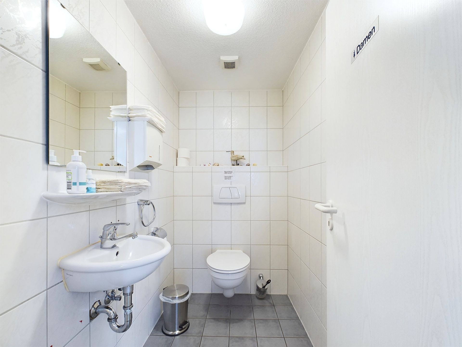 Helles Badezimmer mit Waschbecken, Spiegel, WC und weißen Fliesen an Wand und Boden.