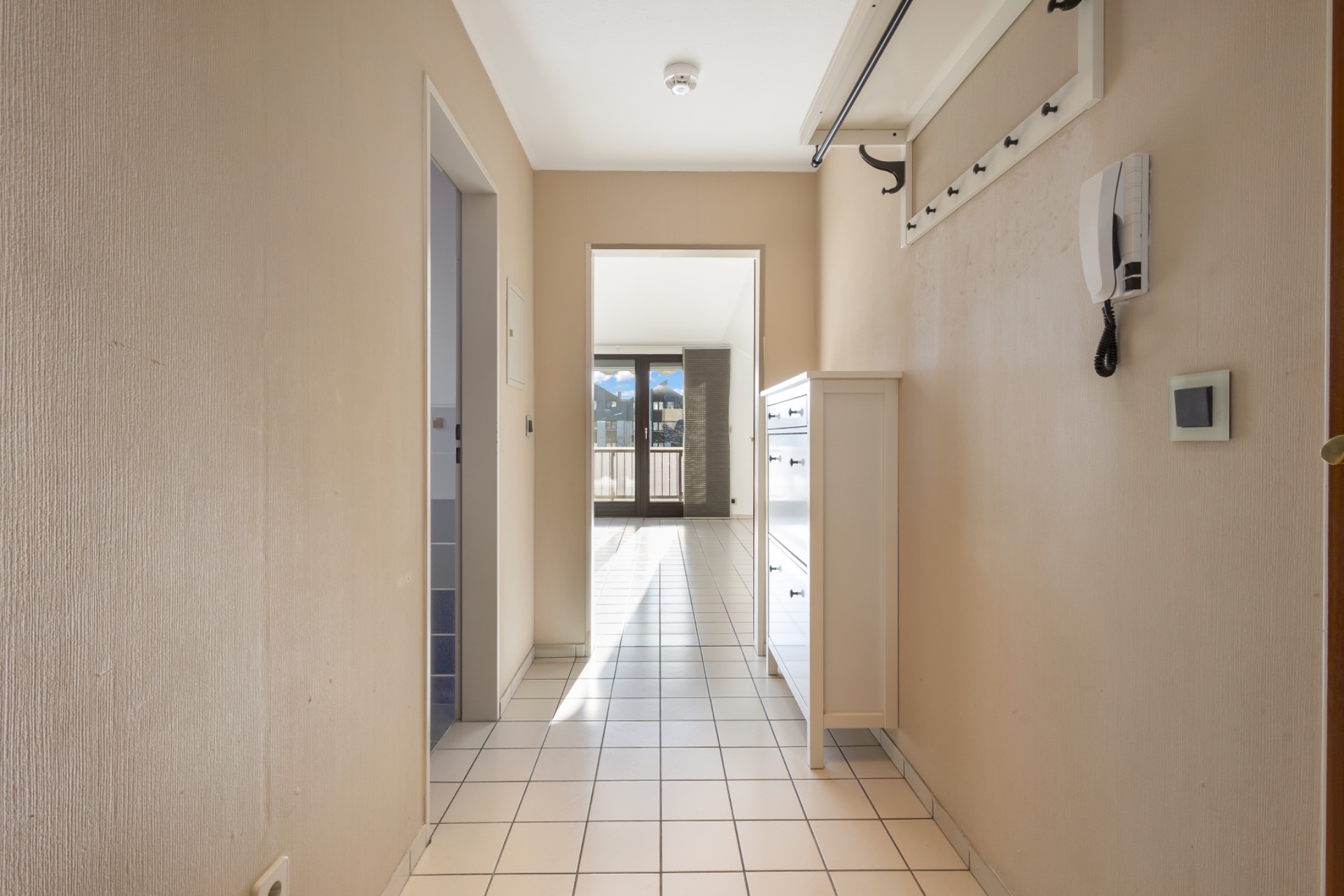 Heller Flur mit weißen Fliesen, Kommode, Garderobe und Blick auf Balkon.
