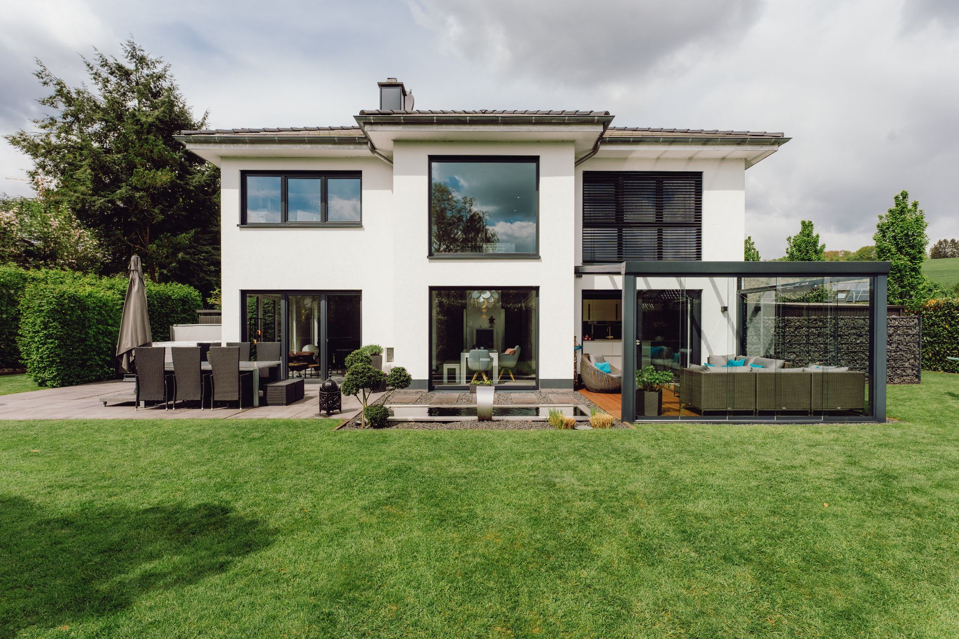 Modernes zweistöckiges Wohnhaus mit Garten, Terrasse und Wintergarten, aufgenommen bei Tageslicht.