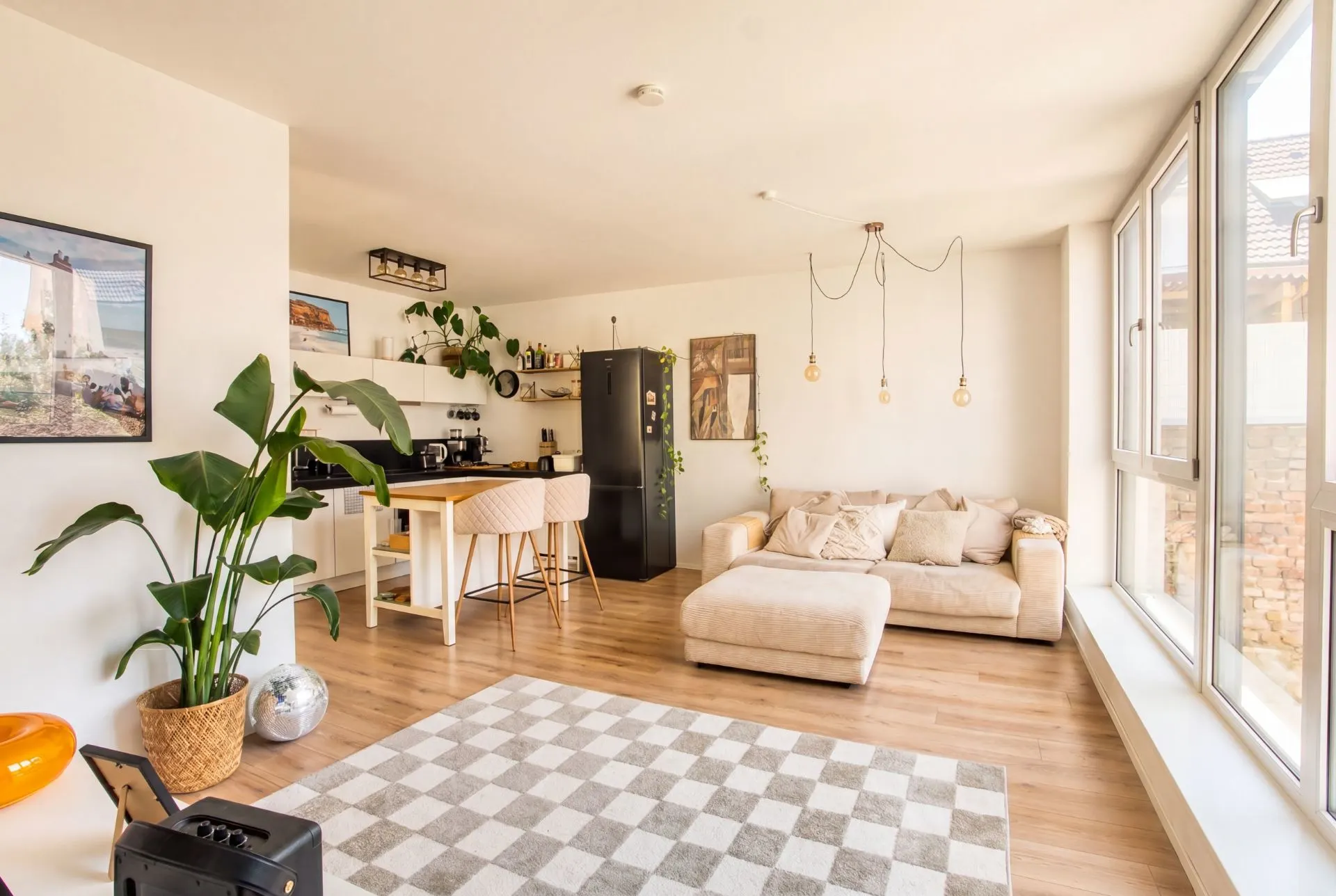 Helles Wohnzimmer mit großer Fensterfront, Sofa, Teppich und offener Küche mit Barhockern.