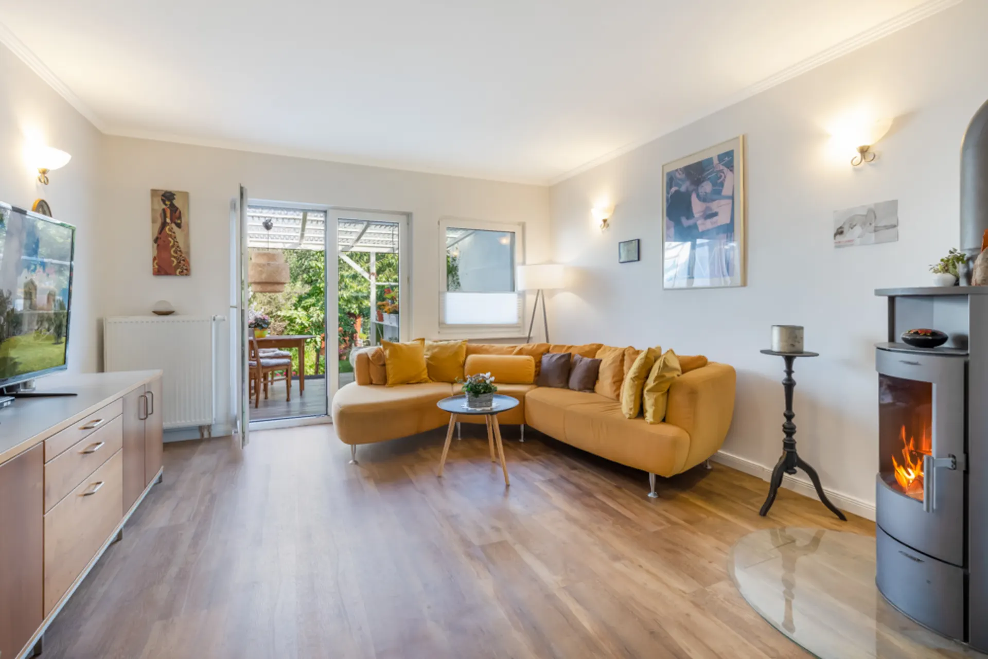 Helles Wohnzimmer mit gelbem Sofa, Holzofen, Fernseher und Zugang zur Terrasse mit Gartenblick.