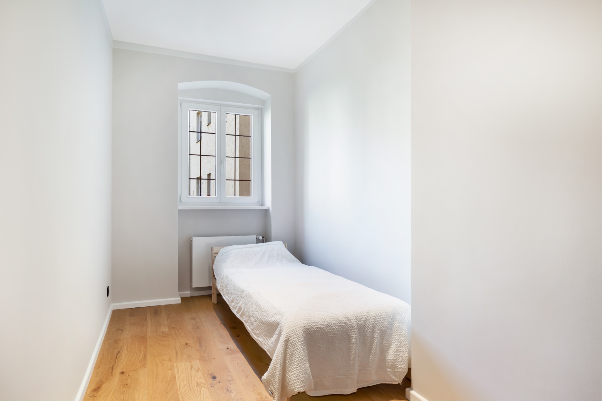 Schlichtes, schmales Schlafzimmer mit Einzelbett, Holzboden und Fenster mit Gitter.