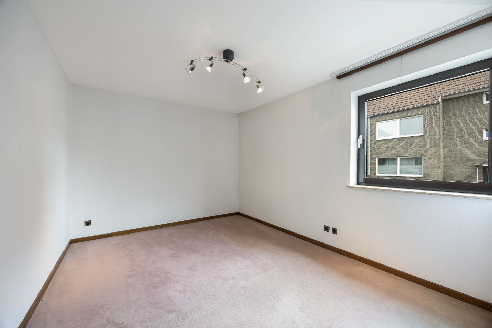 Leeres Zimmer mit Teppichboden, weiß gestrichenen Wänden und großem Fenster.