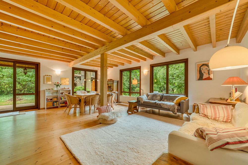 Helles Wohnzimmer mit Holzbalkendecke, großen Fenstern, Esstisch und gemütlichen Sofas.