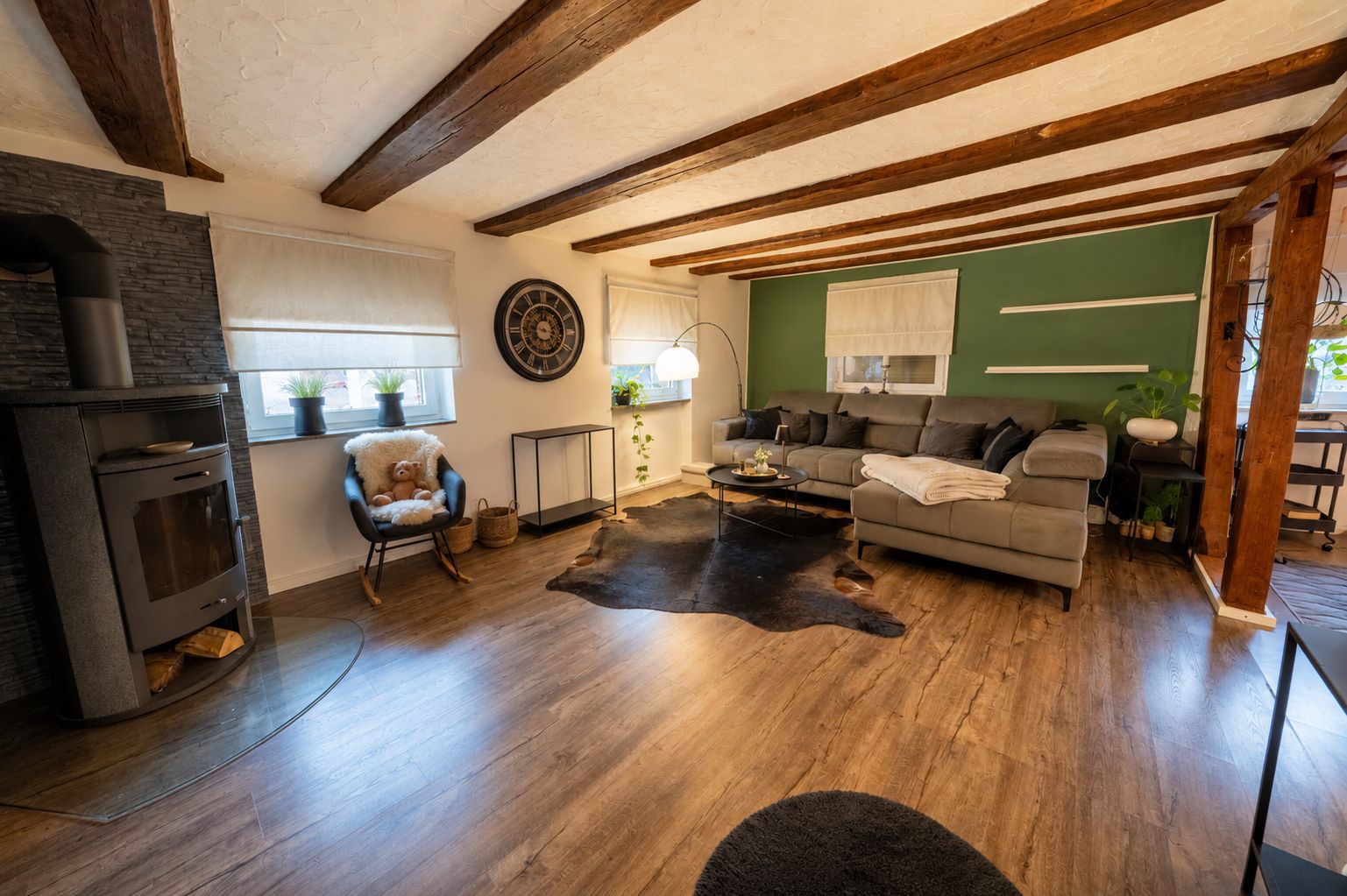 Wohnzimmer mit Holzdecke, Kaminofen, grauem Ecksofa, Teppich und Pflanzen auf Holzfußboden.