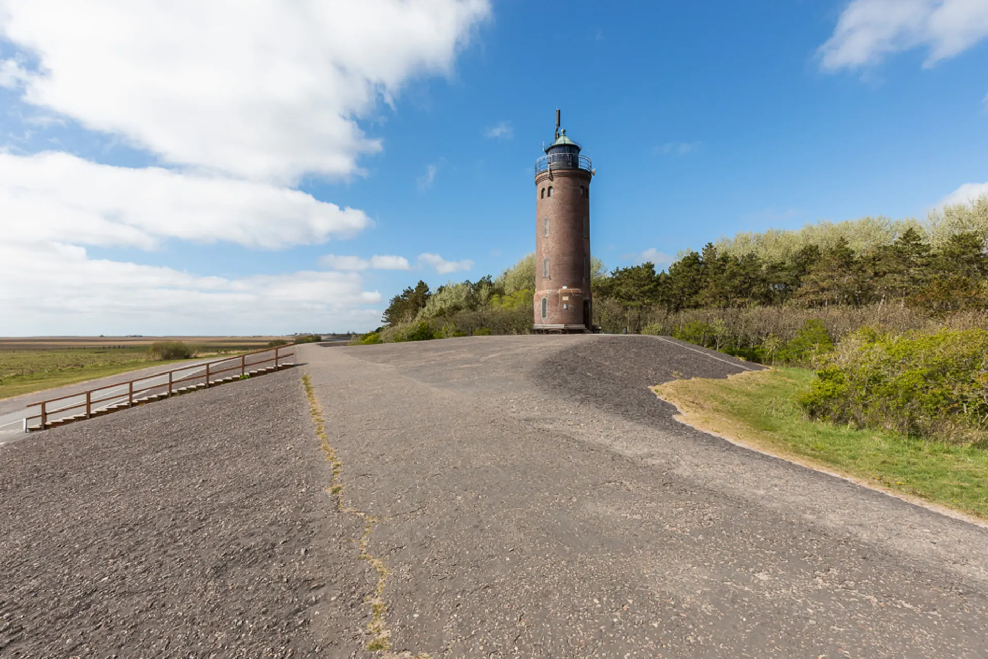 Backsteinturm steht auf einem Hügel neben einer Straße, im Hintergrund Bäume und Wiese.