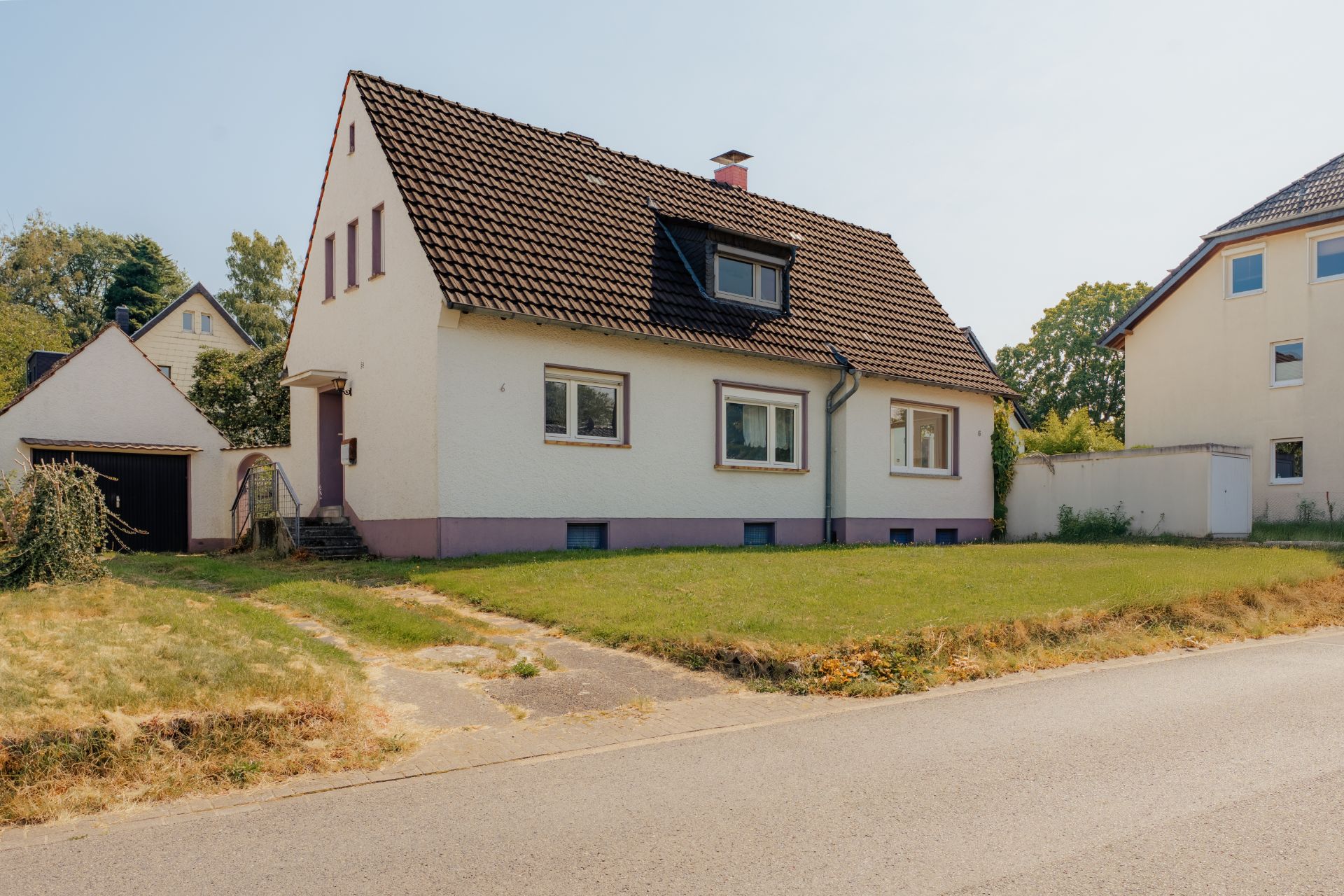 Weißes Einfamilienhaus mit braunem Dach und Vorgarten an einer Straße.