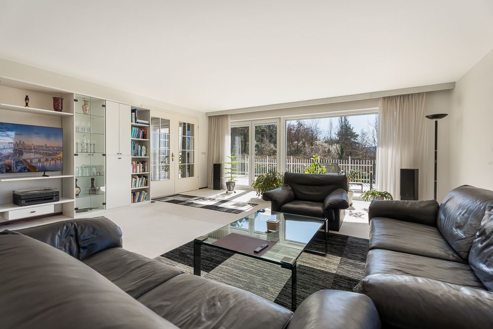 Helles Wohnzimmer mit Ledersofas, Glastisch, Regalwand und großen Fenstern mit Blick nach draußen.