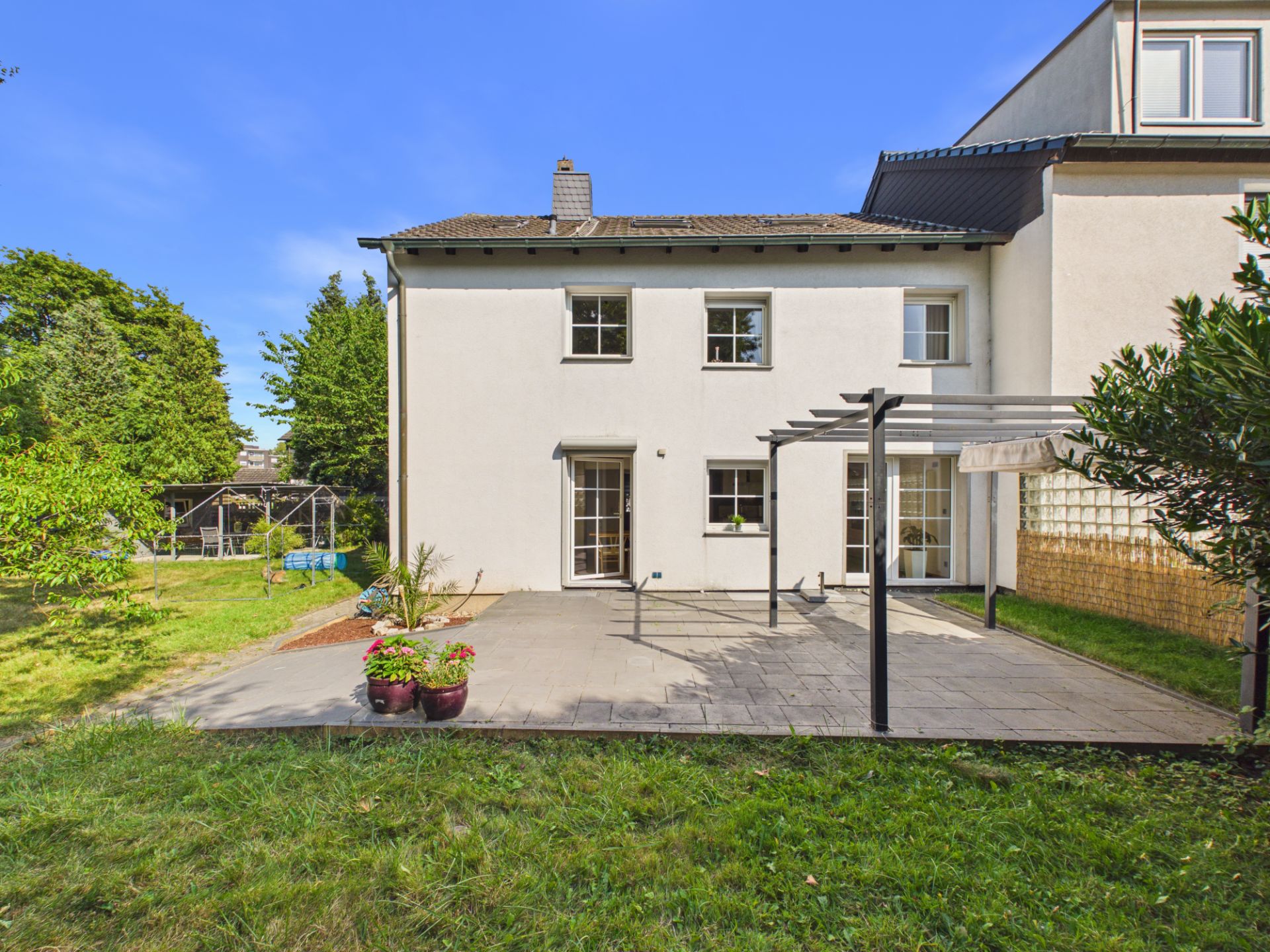 Weiße Hausrückseite mit Terrasse, Rasenfläche und mehreren Pflanzkübeln im Garten.