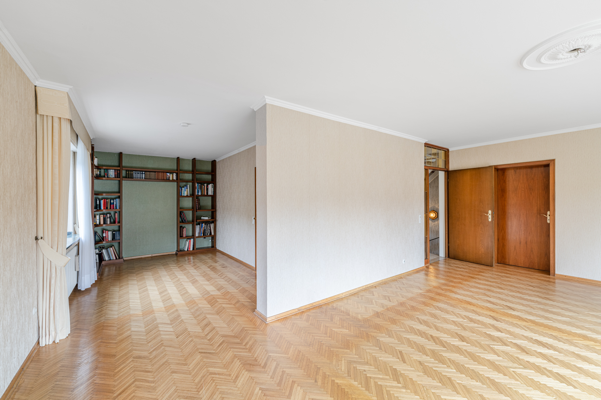 Helles Wohnzimmer mit Parkettboden, Bücherregal, zwei Holztüren und großen Fenstern mit Vorhängen.