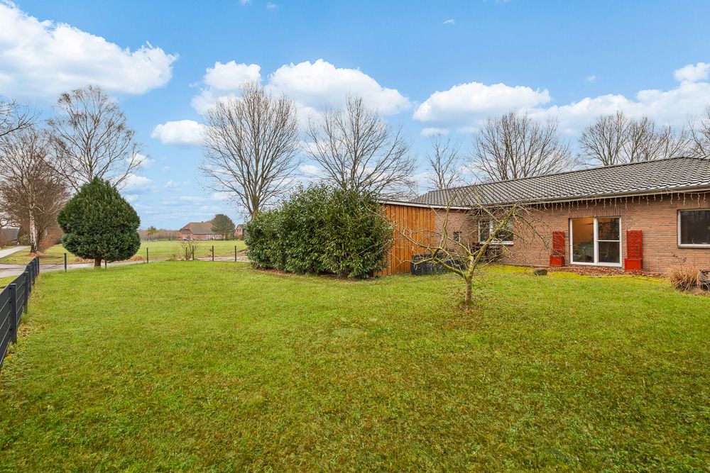 Garten mit Rasen, Büschen, Baum und Haus mit rotem Fensterladen im Hintergrund.