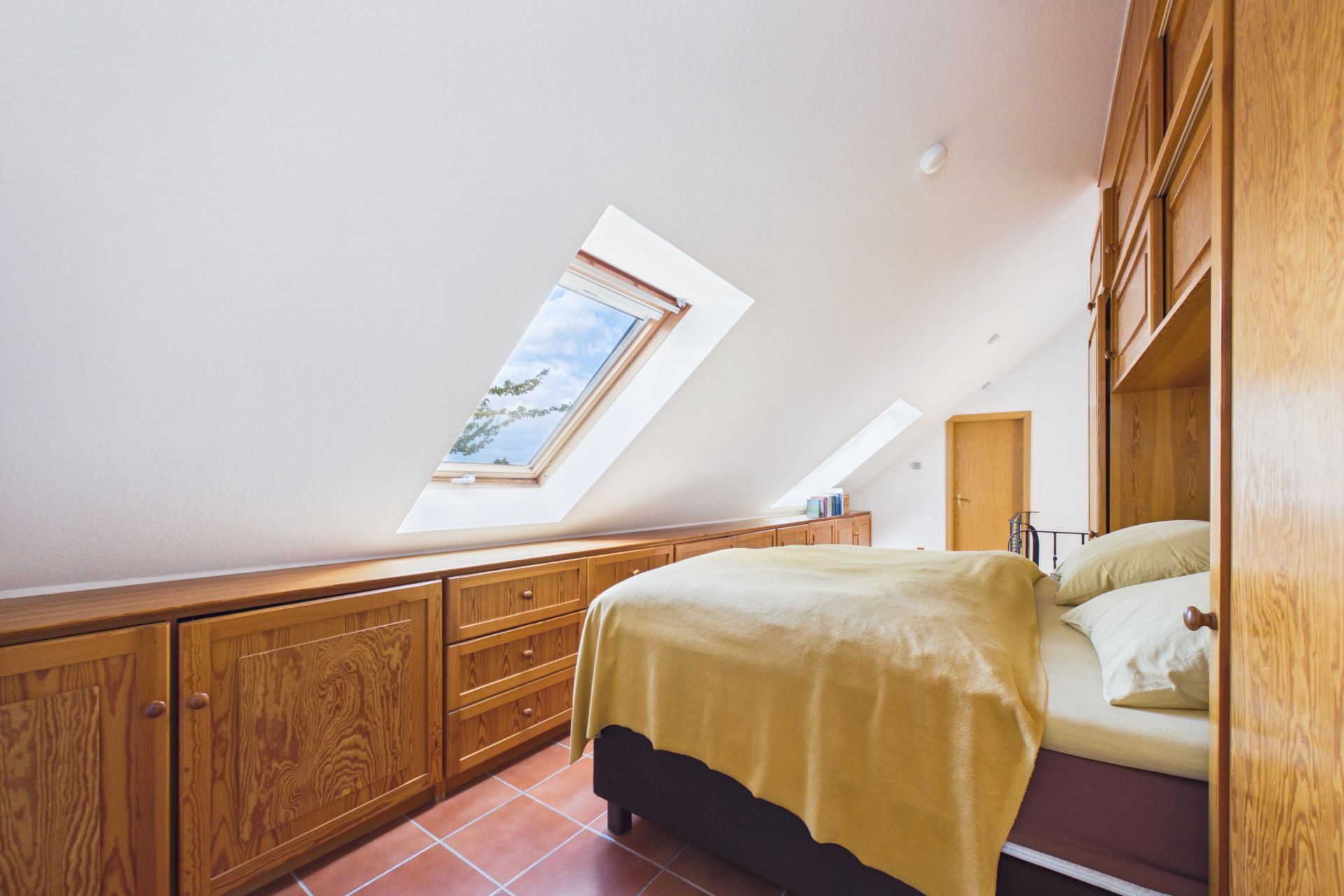 Schlafzimmer mit Dachschräge, Dachfenster, Holzmöbeln und Bett mit gelber Decke.
