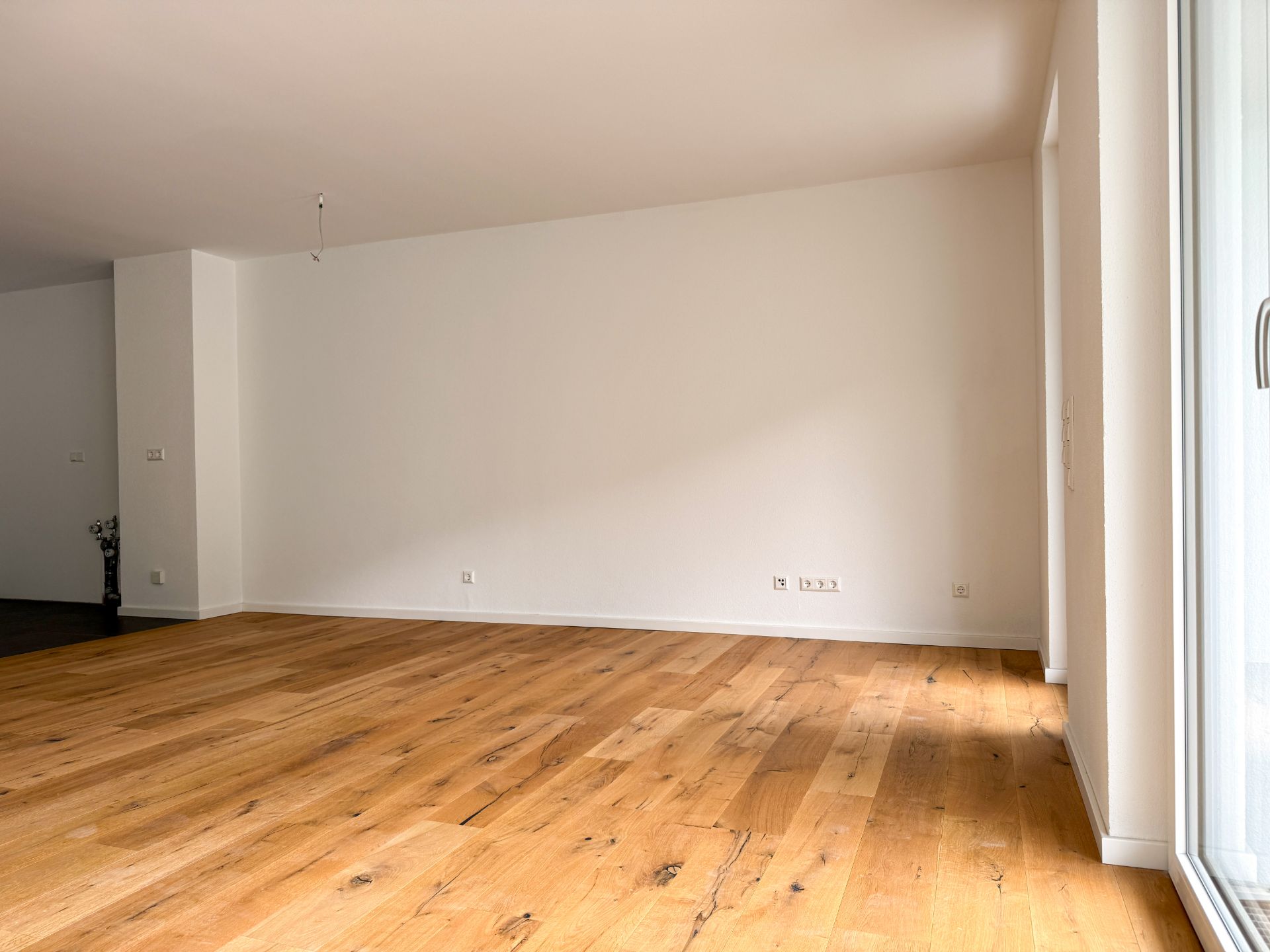 Leerer Raum mit Holzfußboden, weißen Wänden und einem großen Fenster rechts.