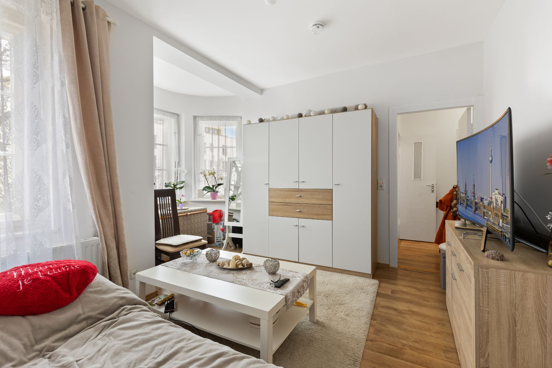 Helles Wohnzimmer mit Sofa, Couchtisch, Schrank, Fernseher und großem Fenster.