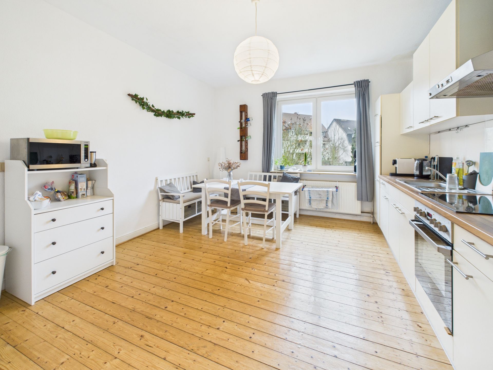 Helle Küche mit Holzboden, weißem Esstisch, Stühlen und großem Fenster mit Vorhängen.