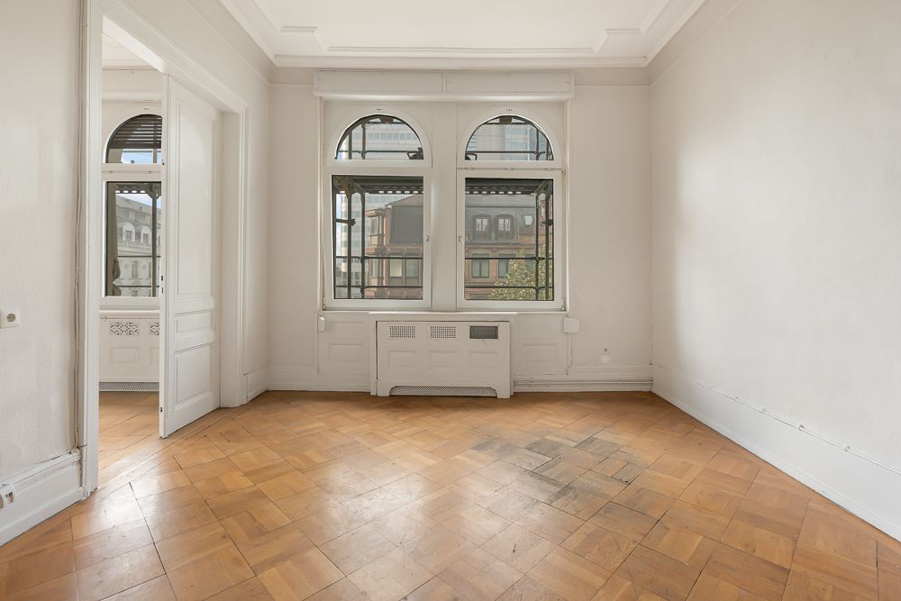 Leerer Raum mit Parkettboden, zwei großen Fenstern und weißen Wänden.
