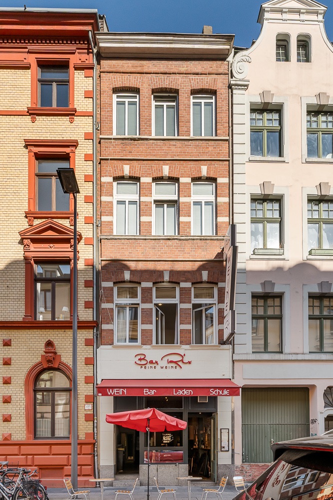 Mehrstöckiges Wohnhaus mit roter Markise und Tischen vor dem Eingang.