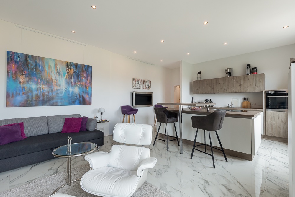 Modernes Wohnzimmer mit offener Küche, grauem Sofa, weißem Sessel und Marmorboden.