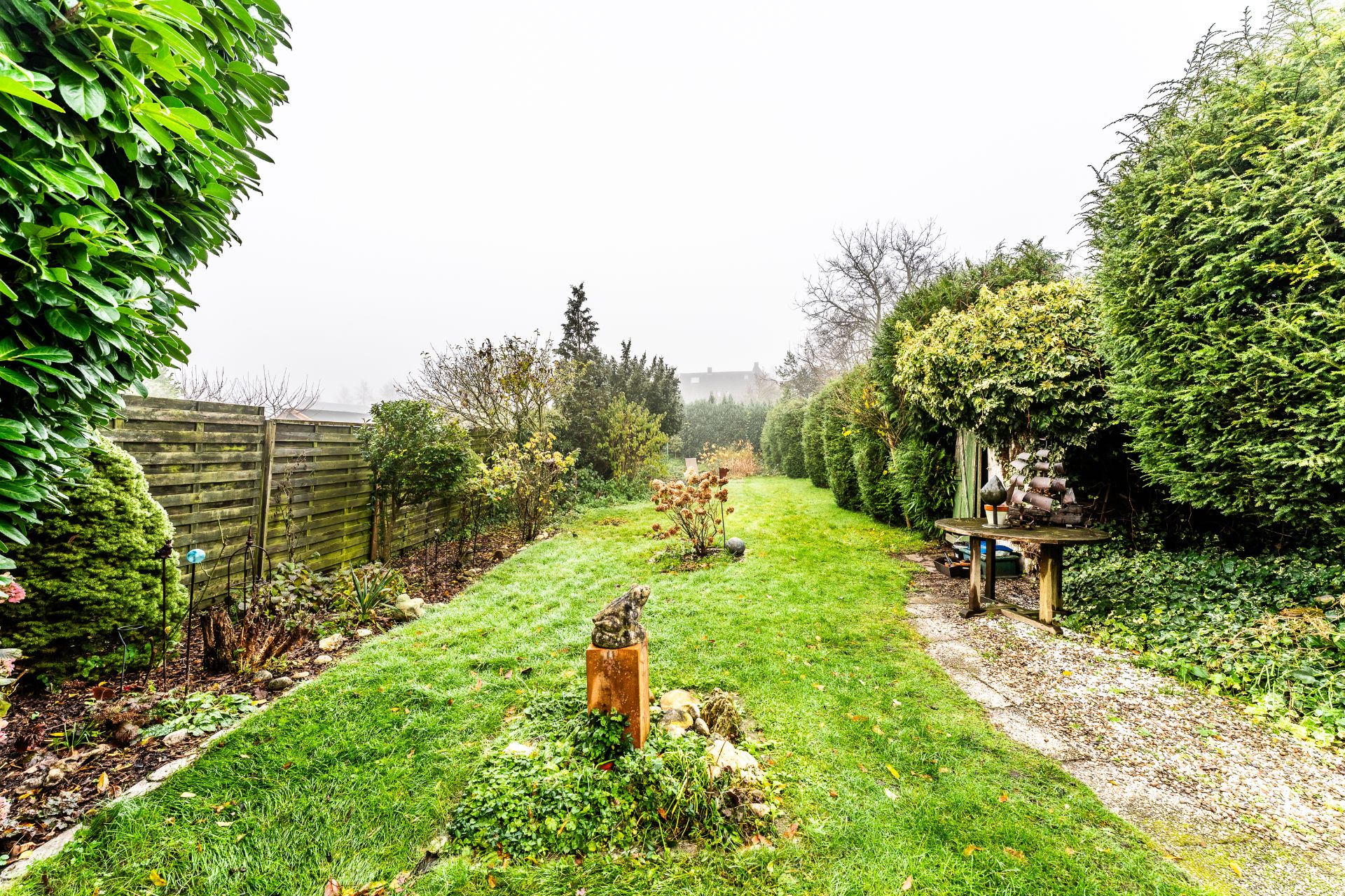Langer Garten mit Rasen, Hecken und einem Holztisch rechts neben einem Kiesweg.