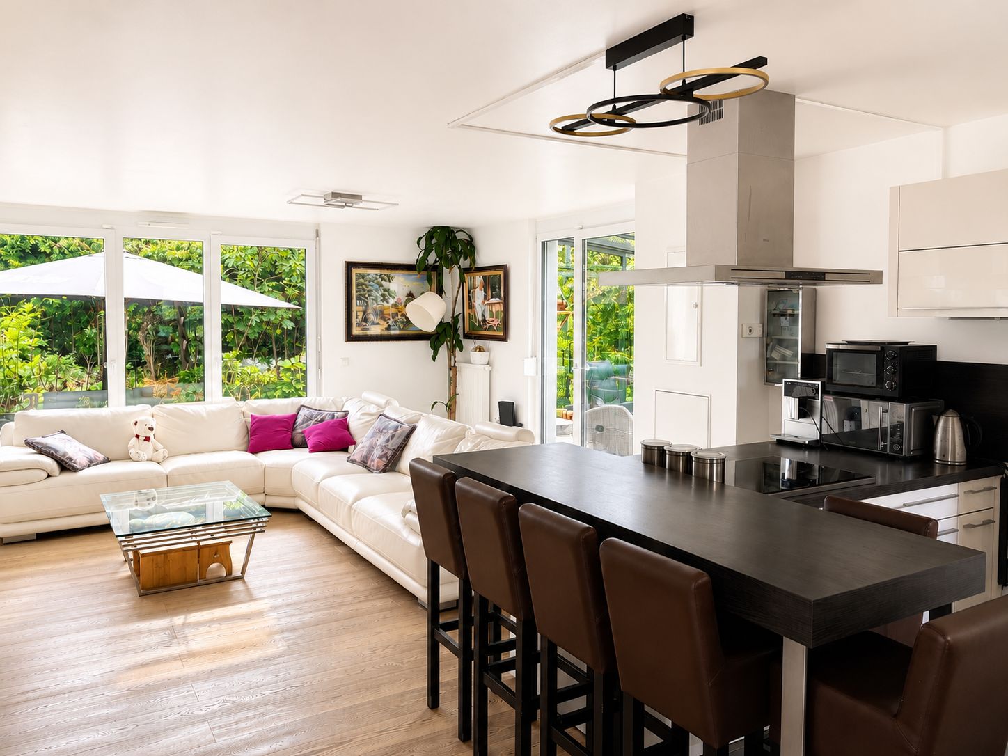 Modernes Wohnzimmer mit großer Eckcouch, Kücheninsel und Blick auf den Garten durch große Fenster.