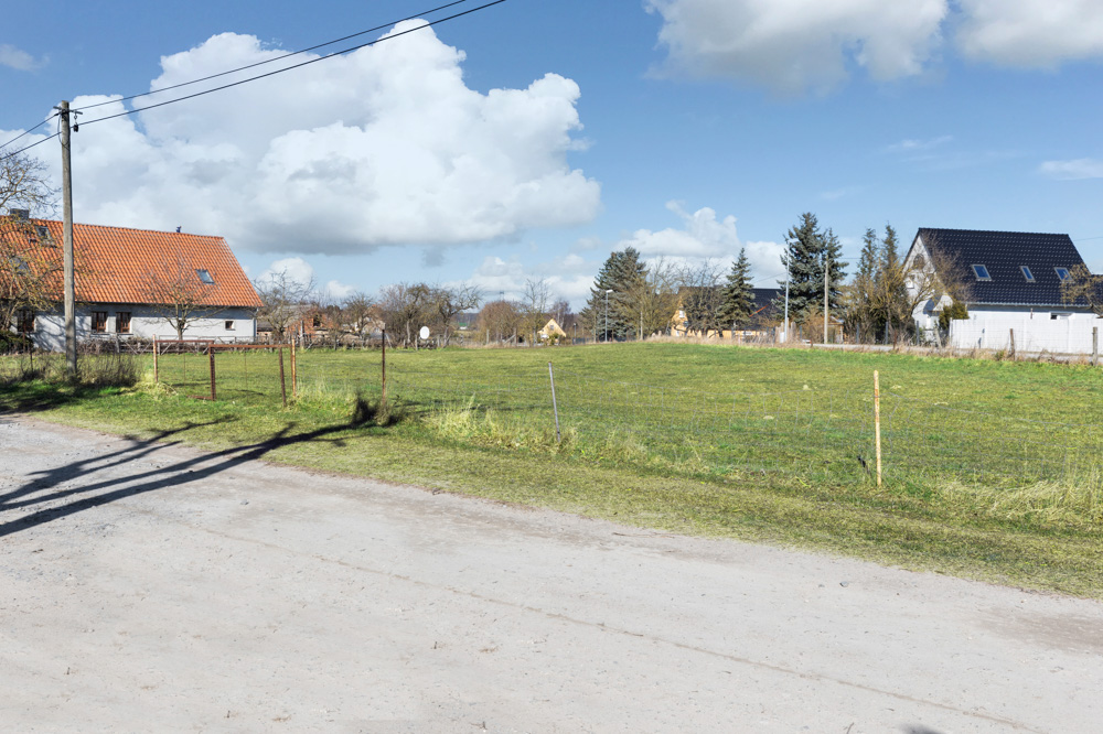 Grünes, leeres Grundstück an einer unbefestigten Straße, mit Häusern im Hintergrund.