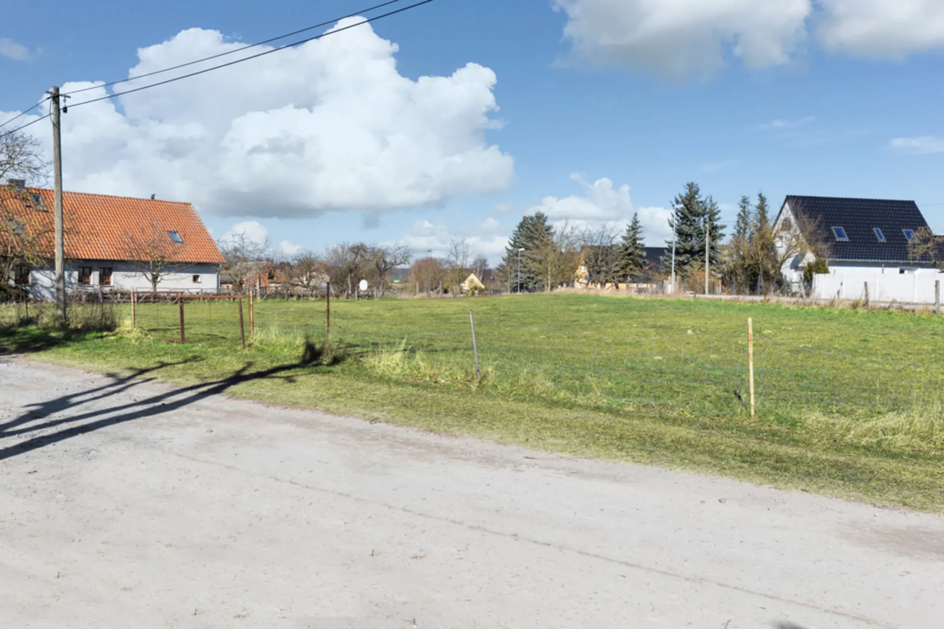 Grünes, leeres Grundstück an einer unbefestigten Straße, mit Häusern im Hintergrund.