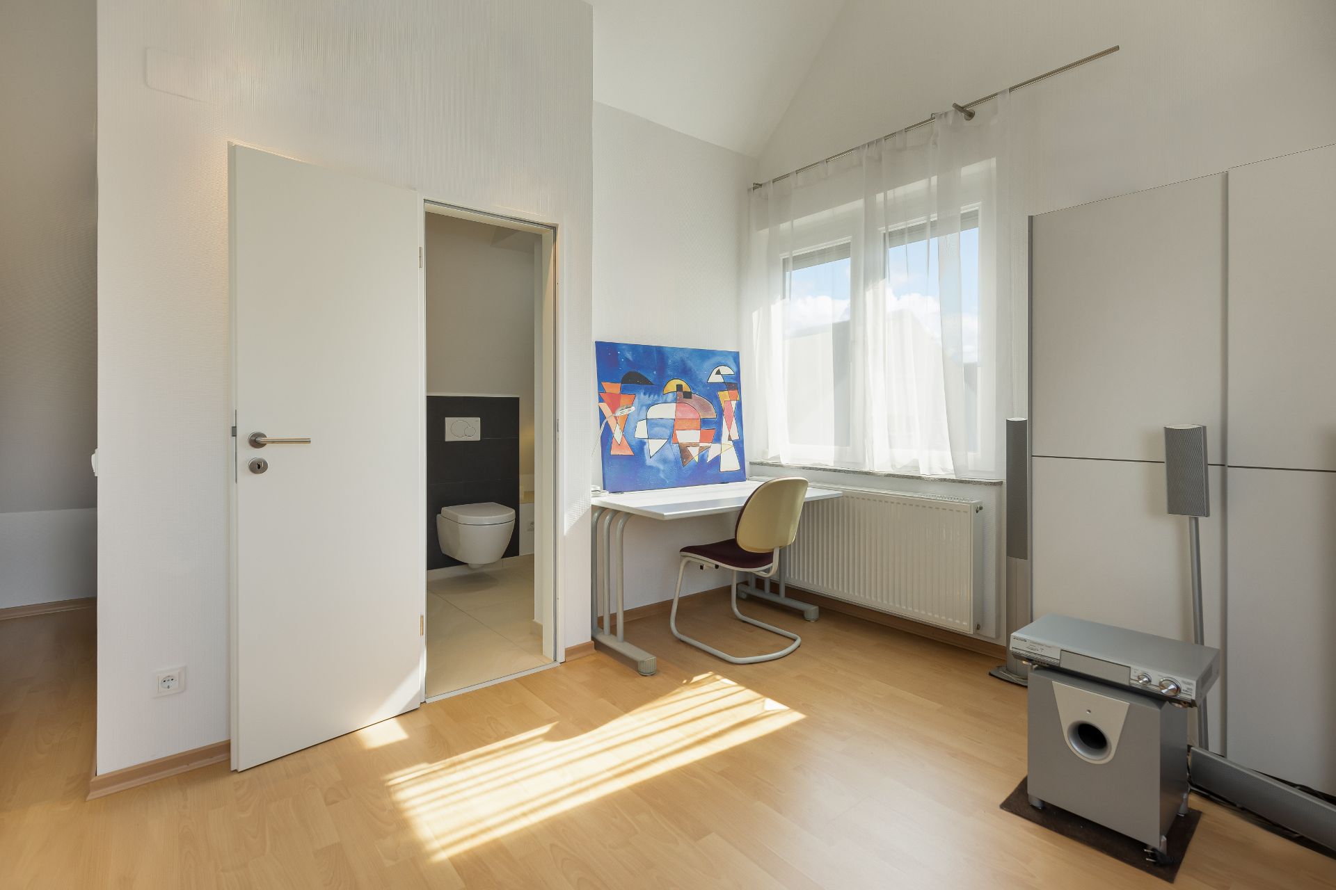 Helles Zimmer mit Schreibtisch, Stuhl, Fenster, Kleiderschrank und offenem Zugang zu einem Bad mit Toilette.