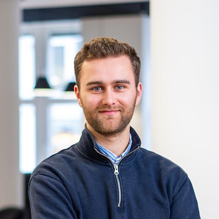 Ein junger Mann mit Bart steht mit verschränkten Armen in einem hellen Raum.