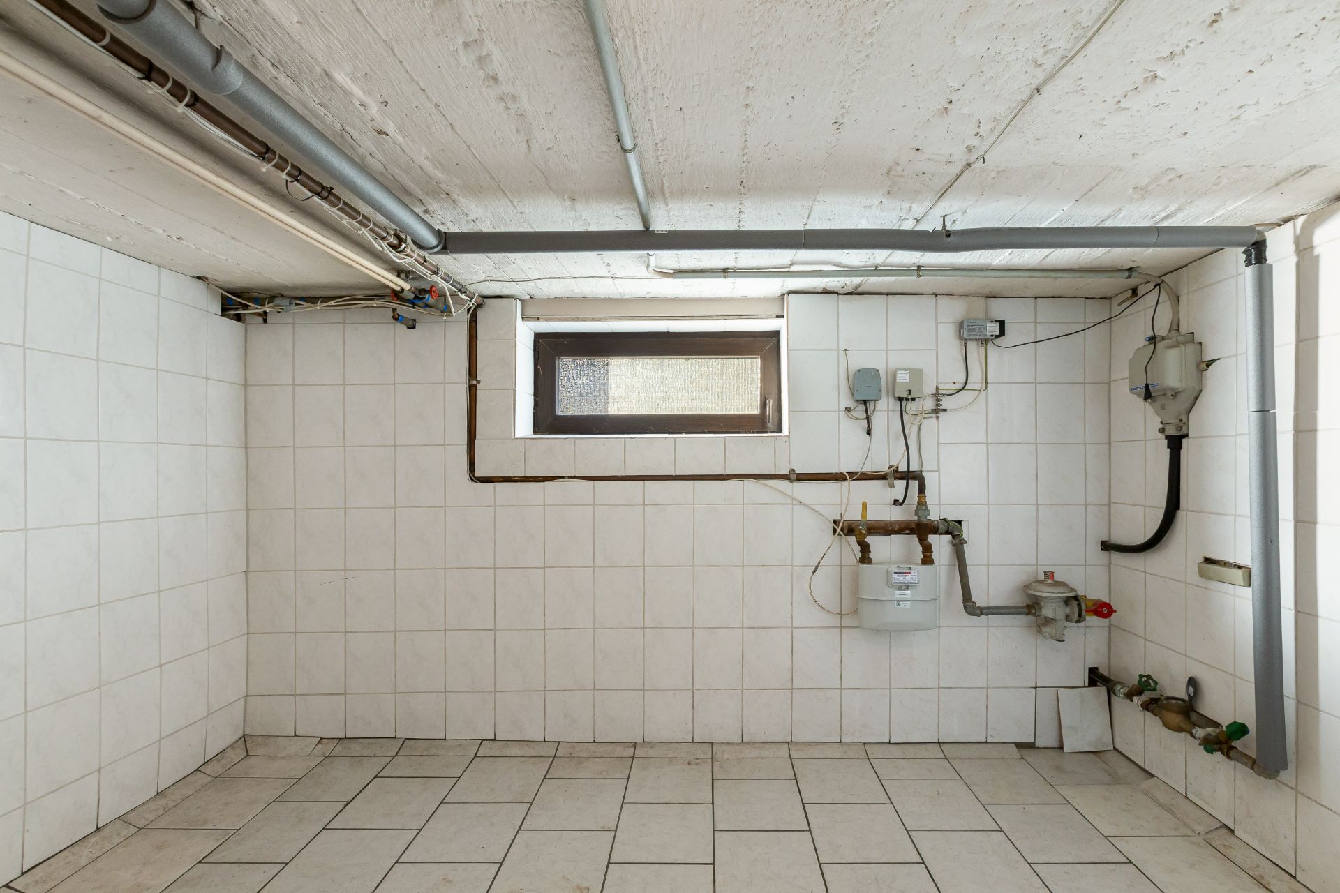 Gefliester Kellerraum mit kleinen Fenstern, Rohren und Hausanschlüssen an der Wand.