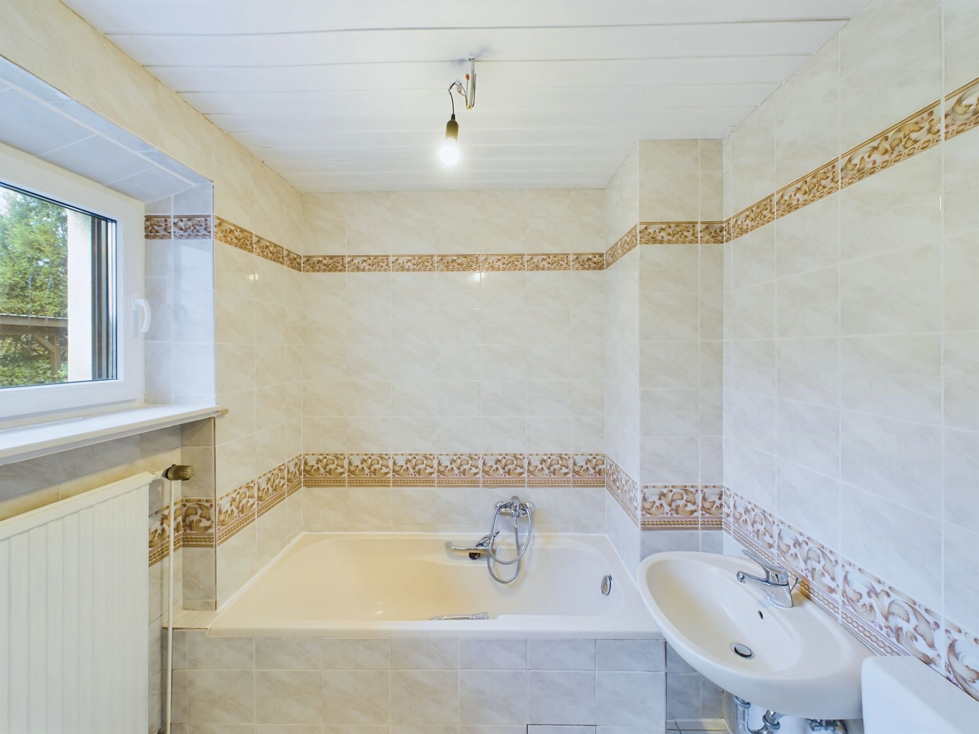 Helles Badezimmer mit Badewanne, Waschbecken, Fenster und gemusterten Fliesen.